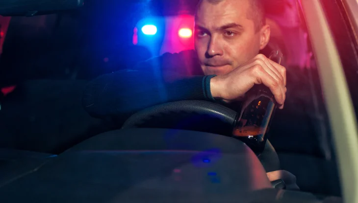 Man drinking behind the wheel with police lights behind him.