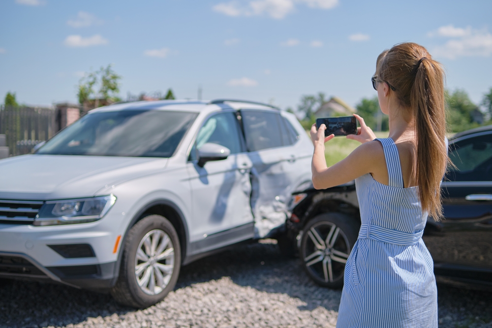 Woman in blue dress taking a photo of a car accident with her cell phone.