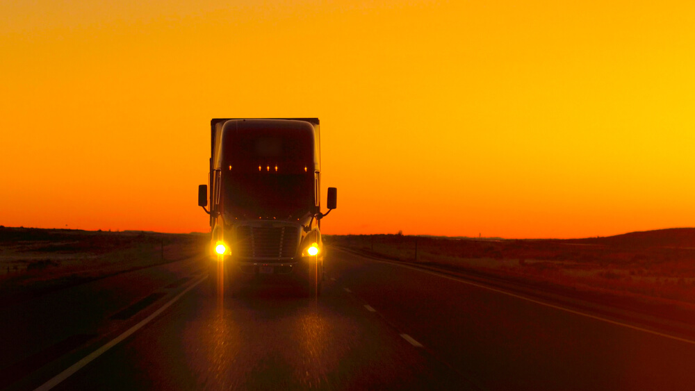 Semi-truck driving with the sunrise behind it, headlights on, cheap commercial truck insurance in Georgia.