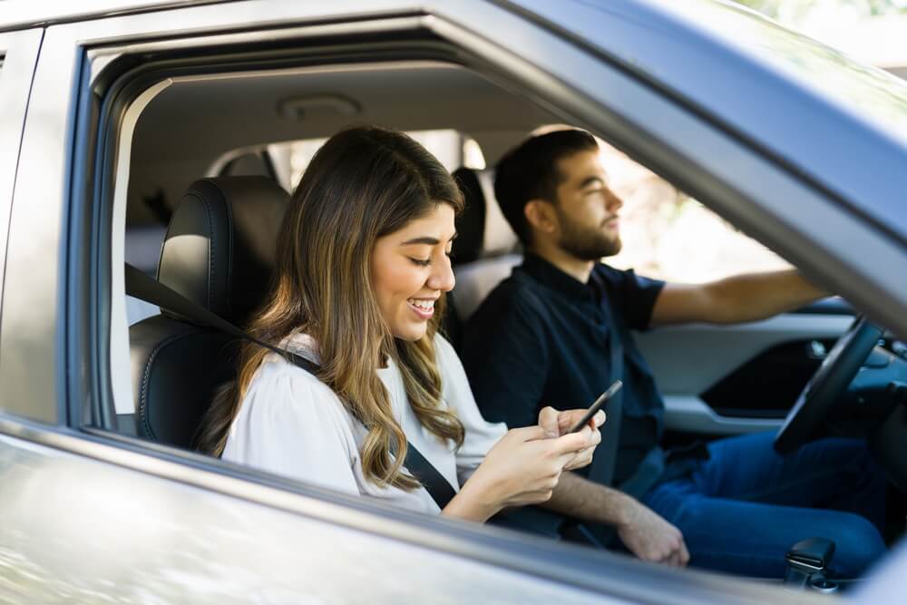 Happy couple driving with man paying attention and woman is passenger on phone - cheap car insurance in Georgia.