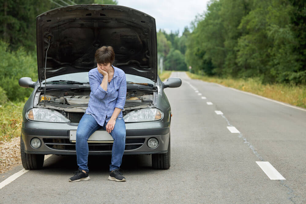 Worried woman sits on the bumper of her broken car on a rural road