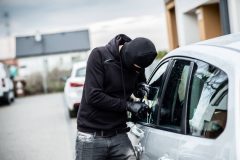 Masked man in a hoodie breaking into a car with a tool.