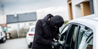 Masked man in a hoodie breaking into a car with a tool.