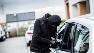 Masked man in a hoodie breaking into a car with a tool.