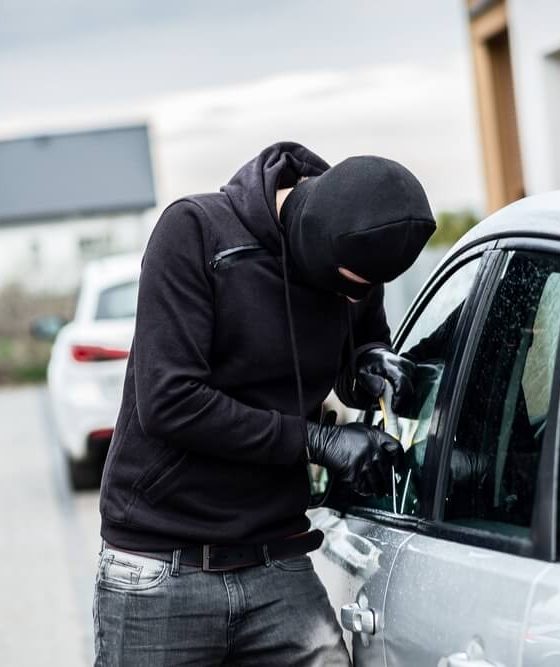 Masked man in a hoodie breaking into a car with a tool.