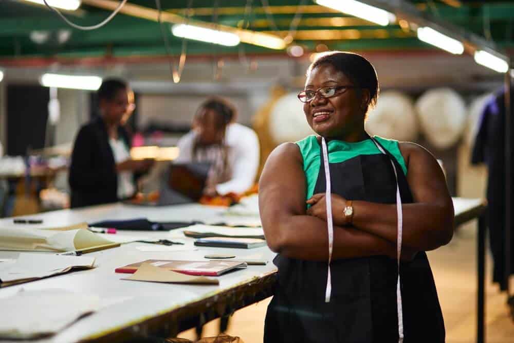 Black woman business owner arms crossed with tape measure around her neck, apron on and arms crossed.