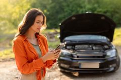 Brunette woman in orange shirt frustrated in front of broken down car calling someone.