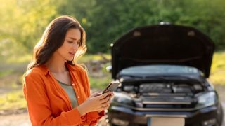 Brunette woman in orange shirt frustrated in front of broken down car calling someone.