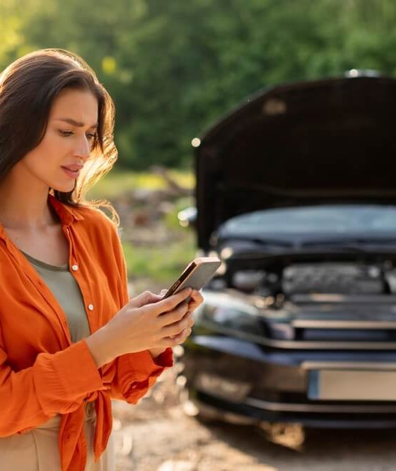 Brunette woman in orange shirt frustrated in front of broken down car calling someone.