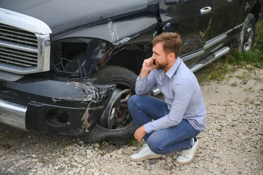 A man squats down to examine the damage to his car after an accident.