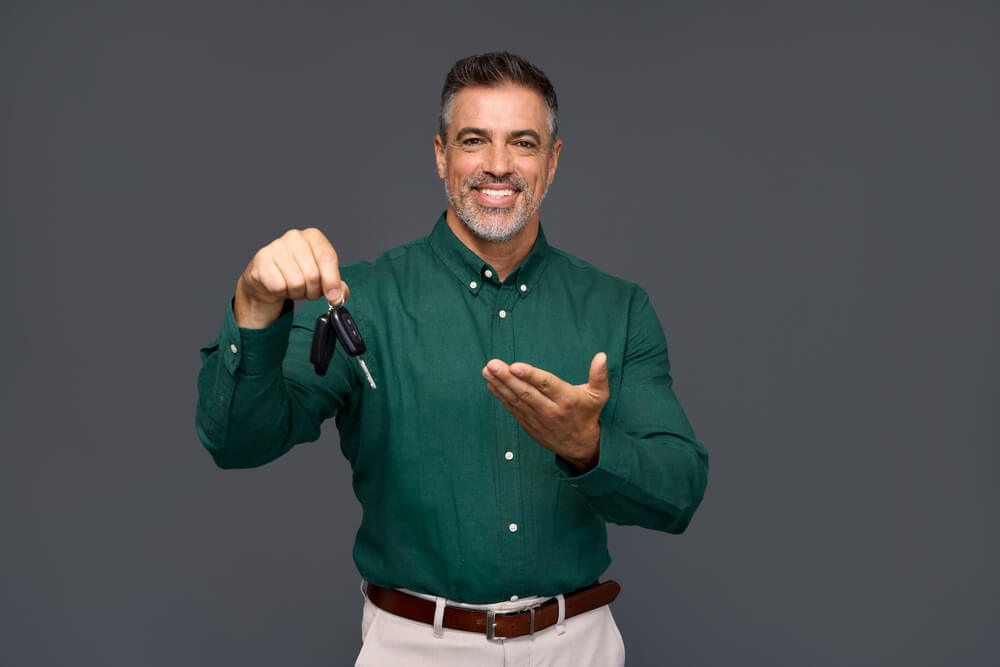 A man in a green shirt stands smiling and holding out the keys to a car he has for sale in Georgia.