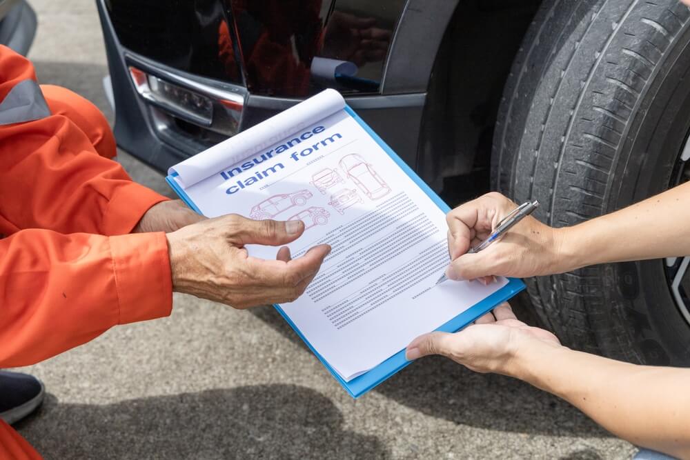 Two people's hands holding and filling out an insurance claim form with a car in the background - cheap car insurance in Georgia.