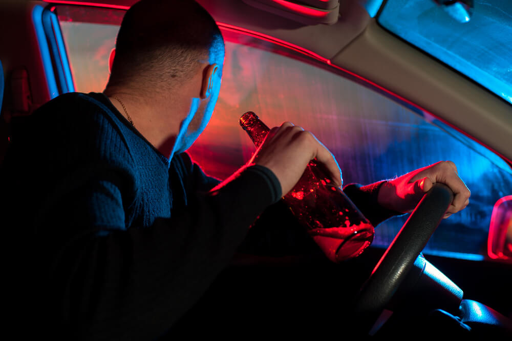 A man behind the wheel holds a bottle of alcohol and looks at red and blue flashing lights, concept o getting arrested for drunk driving - cheap high-risk car insurance in Georgia.