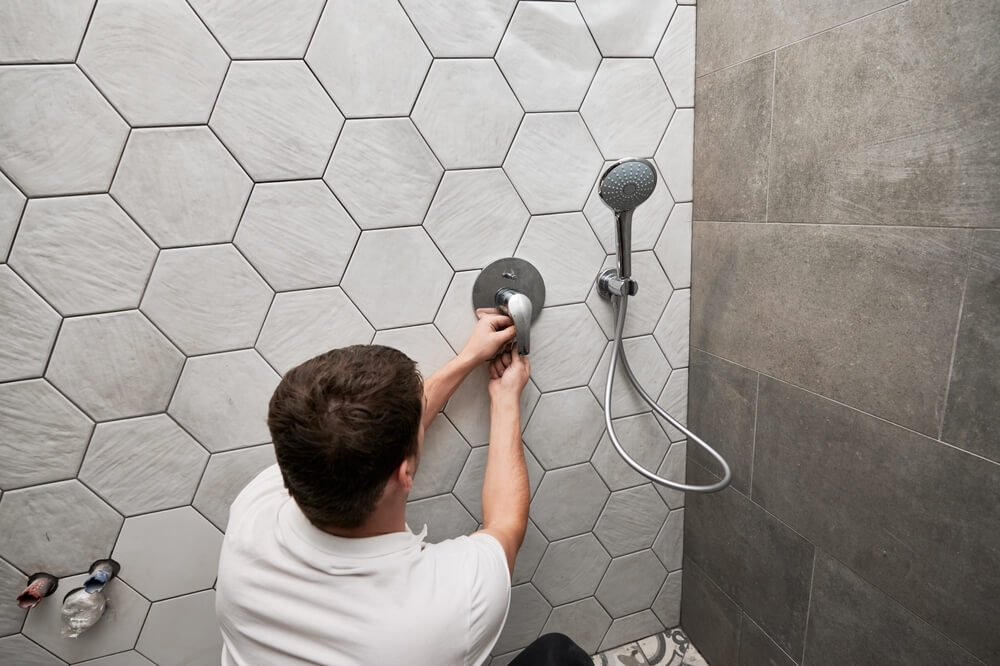 Back view of male installing wall mounted shower system in apartment under renovation. Man repairing metal shower faucet handle while working on bathroom renovation. How renovation affects home insurance.