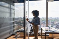 Young executive manager businesswoman writing strategy ideas for her professional commercial business insurance in Georgia on sticky notes on whiteboard doing business project presentation in contemporary corporate office with panoramic view.