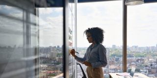 Young executive manager businesswoman writing strategy ideas for her professional commercial business insurance in Georgia on sticky notes on whiteboard doing business project presentation in contemporary corporate office with panoramic view.