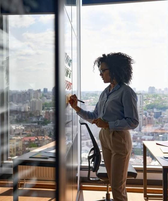 Young executive manager businesswoman writing strategy ideas for her professional commercial business insurance in Georgia on sticky notes on whiteboard doing business project presentation in contemporary corporate office with panoramic view.