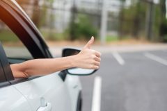 Woman hand thumb ups, switched insurance and got a better price, during driving a car on the road, hand controlling steering wheel in electric modern automobile.
