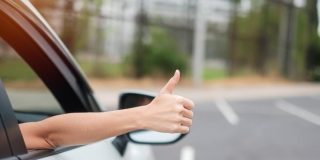 Woman hand thumb ups, switched insurance and got a better price, during driving a car on the road, hand controlling steering wheel in electric modern automobile.