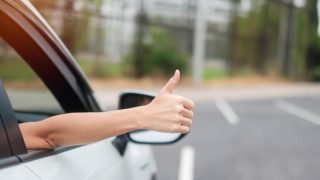 Woman hand thumb ups, switched insurance and got a better price, during driving a car on the road, hand controlling steering wheel in electric modern automobile.