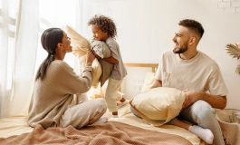 Happy family multiethnic mother, father and son laughing, playing, fights pillows and jumping in bed in bedroom at home, figuring out home insurance deductibles.