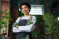 Running of own business. Woman florist wearing apron in botanical store with green plants. Happy small business owner working at flower shop smiling surrounded by plants, protected by cheap business insurance in Georgia.