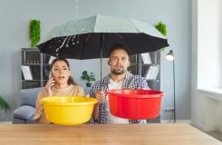 Water leak from ceiling due to roof damage or broken leaky plumbing in house of couple. Woman holding bucket and phone to call repair service, man with umbrella. Emergency, accident at home concept.