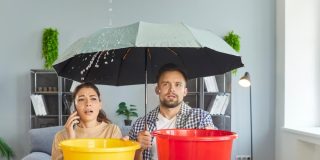 Water leak from ceiling due to roof damage or broken leaky plumbing in house of couple. Woman holding bucket and phone to call repair service, man with umbrella. Emergency, accident at home concept.