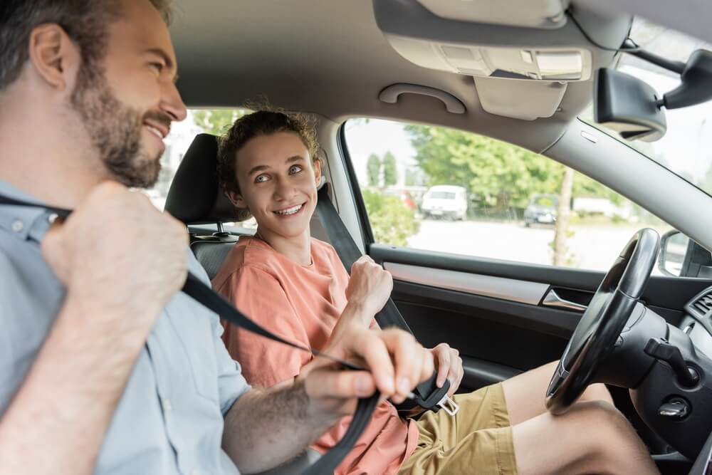 Teen driver smiling at an adult while fastening his seatbelt in the car, highlighting Car Insurance for Teen Drivers during practice driving.