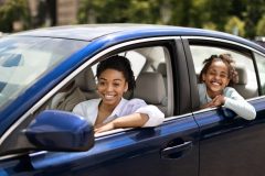 African American Family Posing Sitting In Blue Car In Urban Area. Mom And Daughter Driving New Auto Enjoying Ride Smiling To Camera, avoiding auto insurance mistakes by purchasing coverage.