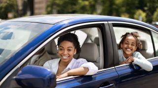 African American Family Posing Sitting In Blue Car In Urban Area. Mom And Daughter Driving New Auto Enjoying Ride Smiling To Camera, avoiding auto insurance mistakes by purchasing coverage.