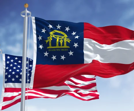 The Georgia state flag waving along with the national flag of the United States of America. In the background there is a clear sky.