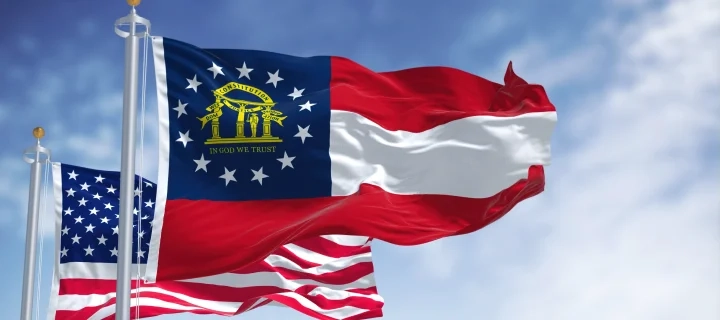 The Georgia state flag waving along with the national flag of the United States of America. In the background there is a clear sky.